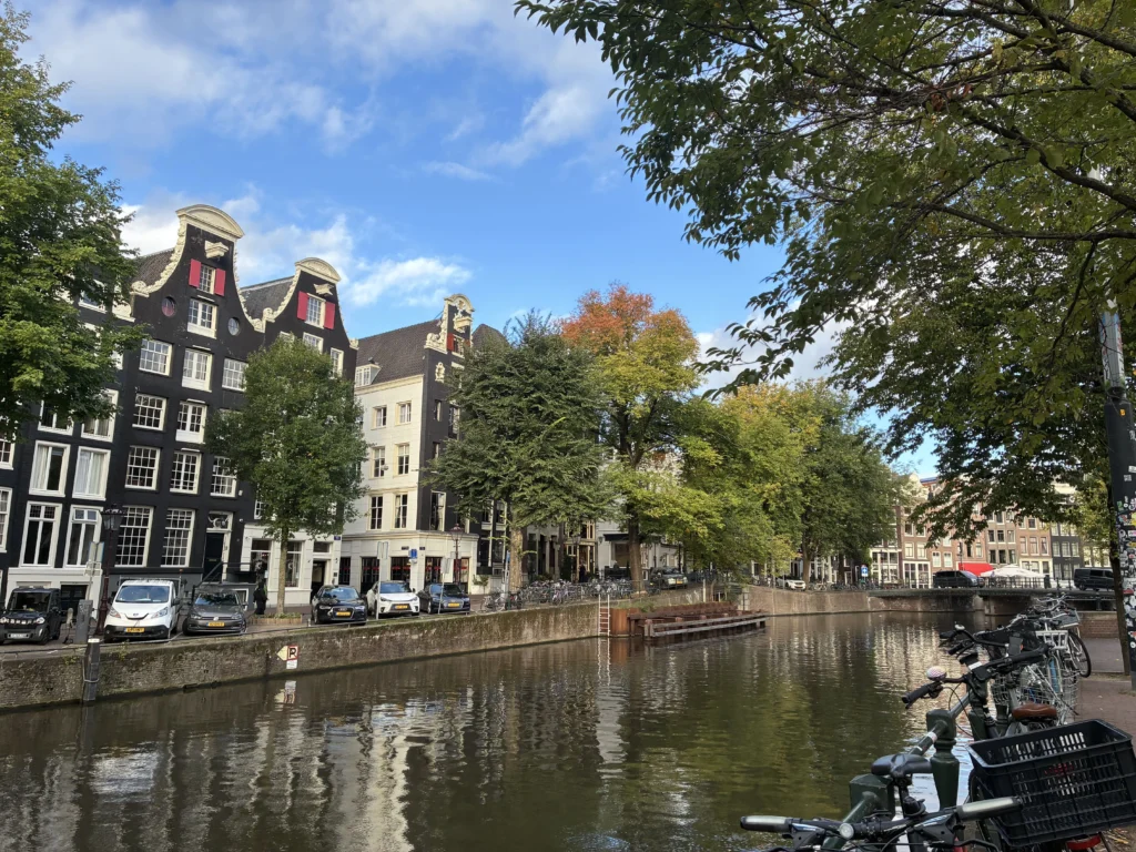 Vista en perspectiva del canal Herengracht en Ámsterdam con fachadas tradicionales holandesas, árboles reflejados en el agua y bicicletas en primer plano.