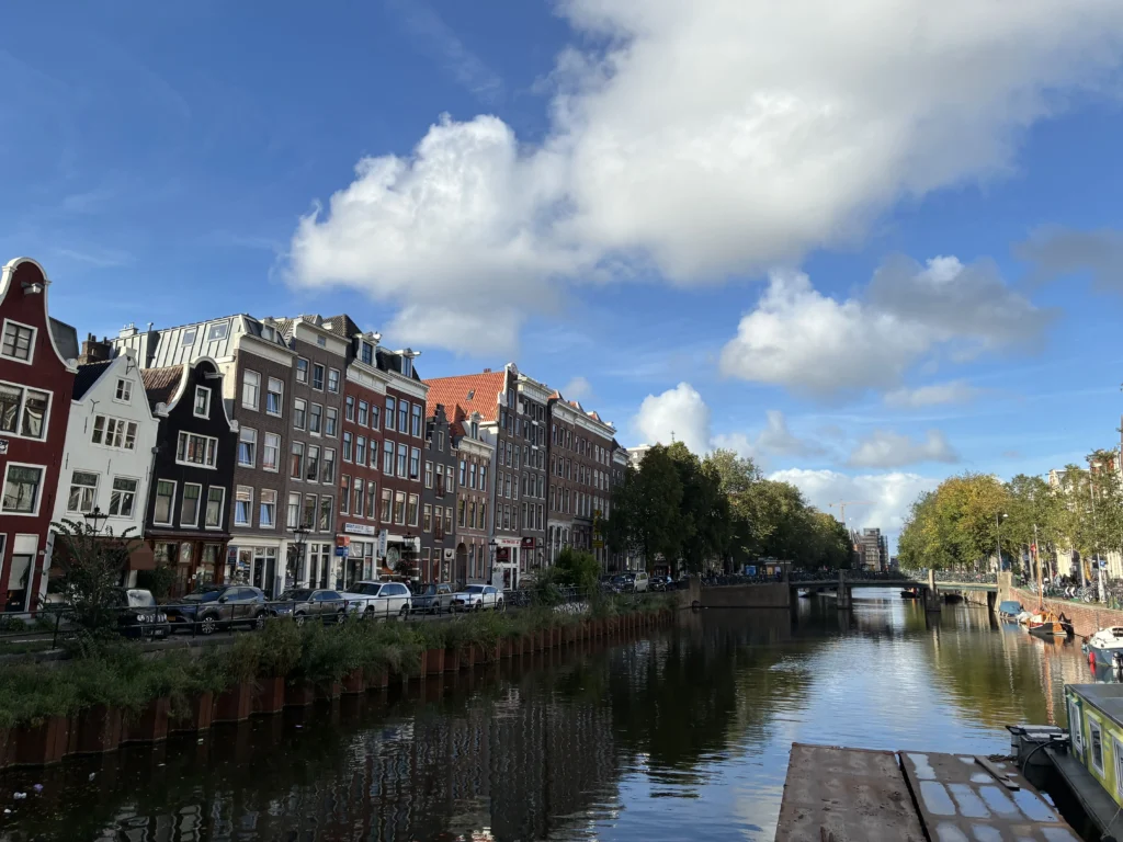 Vista en perspectiva del canal Rokin en Ámsterdam con edificios tradicionales, coches aparcados en el muelle izquierdo y un cielo azul con nubes blancas.
