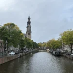 Canal Prinsengracht en Ámsterdam con el canal centrado y la torre de la iglesia Westerkerk destacando a la izquierda bajo un cielo nublado.